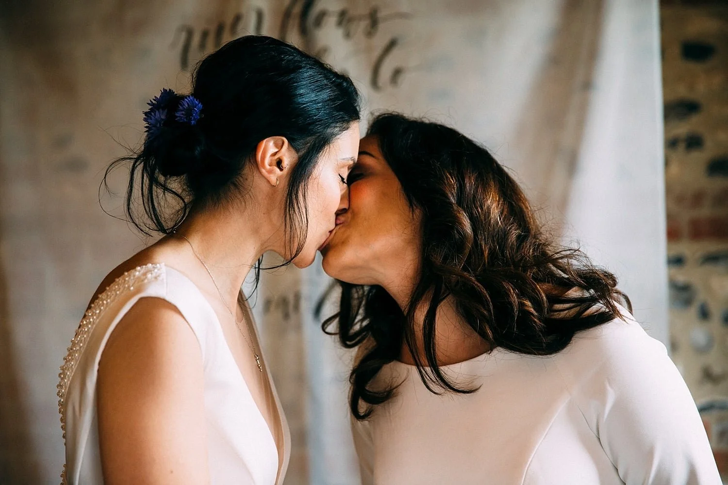 A quiet, intimate moment filled with warmth and tenderness as two brides lean in for a soft, heartfelt kiss. Their foreheads nearly touch, eyes closed, completely wrapped up in each other. One wears a deep-V gown with delicate shoulder beading, her d
