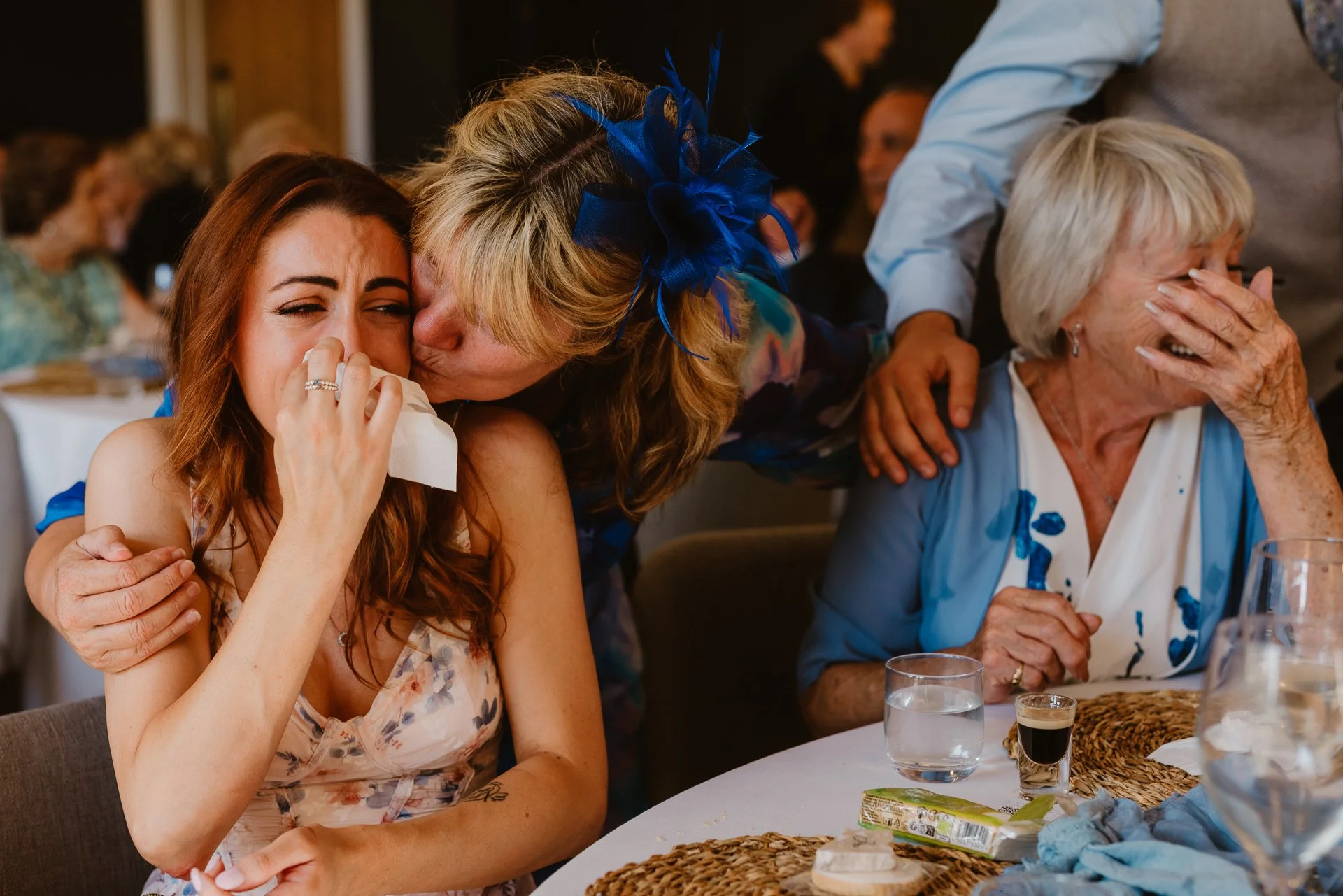 A candid emotional wedding reception moment captured by Bluebell Photo Studio, showing a tearful guest being lovingly hugged and kissed by a family member while another elderly guest laughs beside them. This heartfelt documentary-style wedding photog