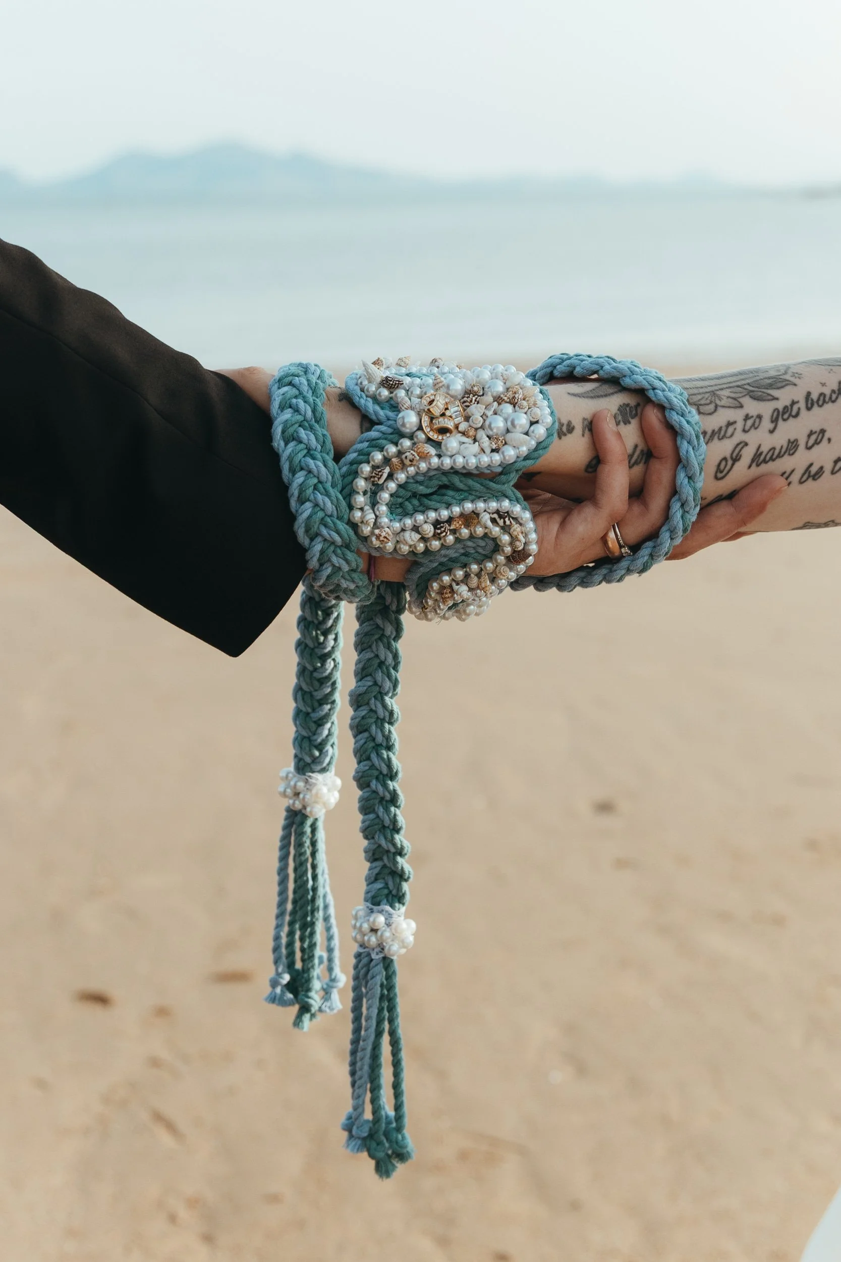 A close-up of two hands bound together with a beautifully crafted hand-fasting cord. The cord is a sea-inspired design in shades of blue and turquoise, adorned with pearls and gold accents, symbolising unity and connection. One person’s hand has tatt