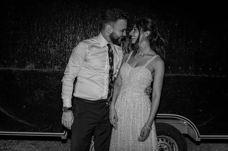 A newly married couple share an intimate moment during their evening celebration, standing close together and smiling softly in a candid black-and-white portrait. The bride’s sparkling gown catches the light as she leans towards her partner, creating