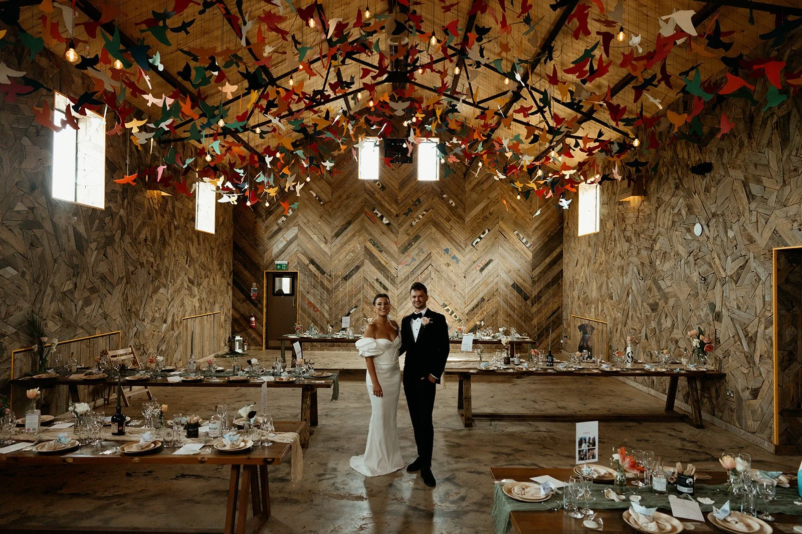 A stylish modern couple couple standing inside The Canary Shed on their wedding day. There is a ceiling full of colourful birds.