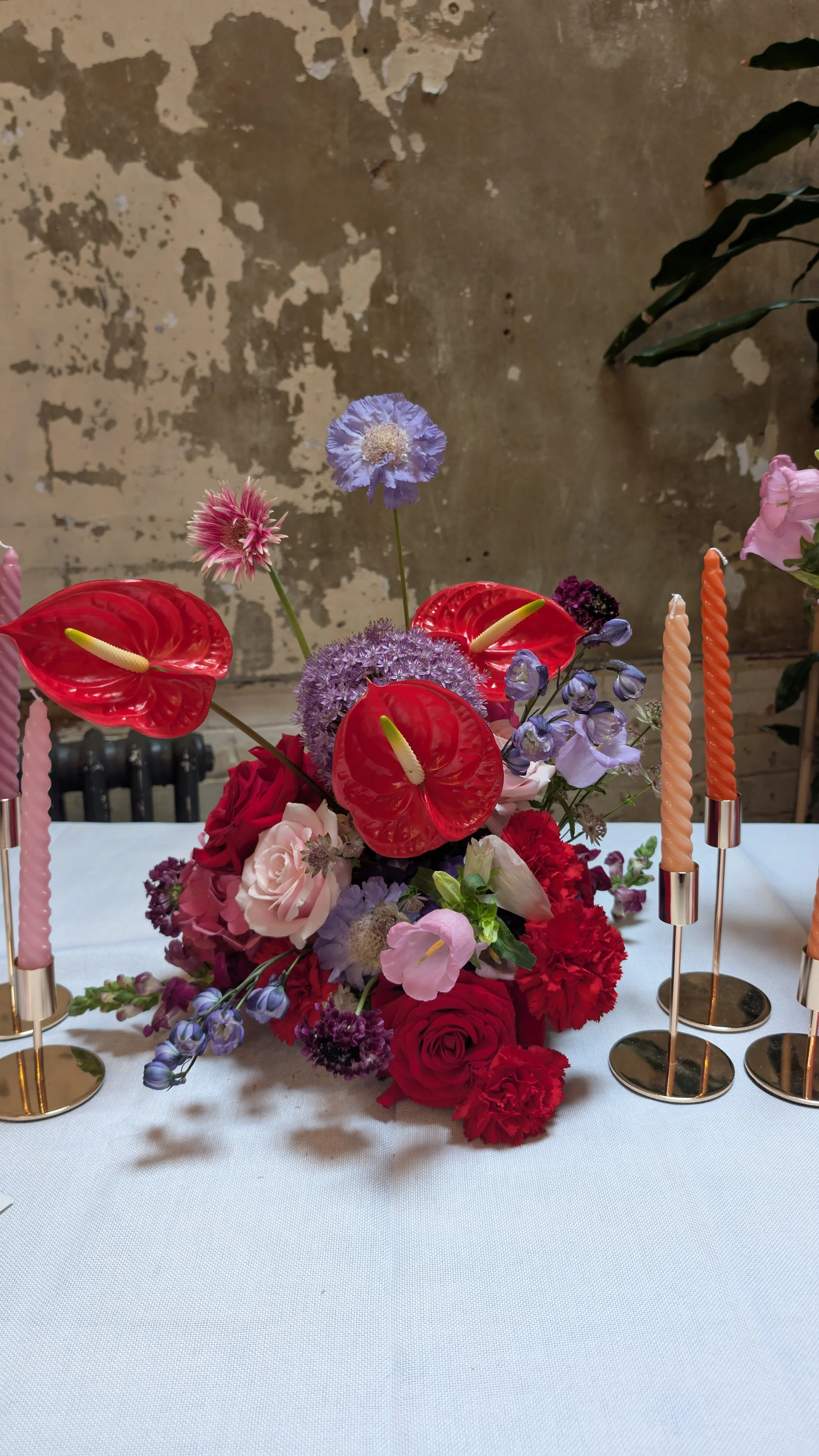 Modern floral centrepiece by Harlequin Florals featuring red anthuriums, pink roses, lilac scabiosa, and purple alliums, styled with pastel twist candles in gold holders. Set against a rustic wall, this bold yet romantic arrangement blends vibrant co