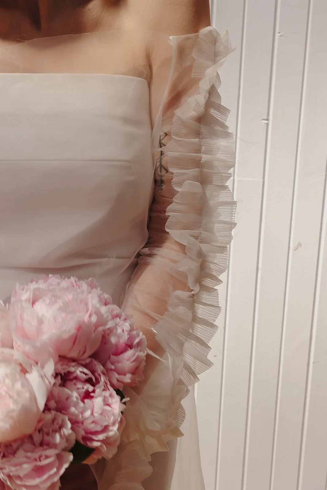 A close-up focuses on the upper body of a bridal look, showing a smooth, strapless bodice paired with a sheer sleeve edged in fine, pleated ruffles. The translucent fabric reveals layered texture and soft movement against the skin, while a cluster of