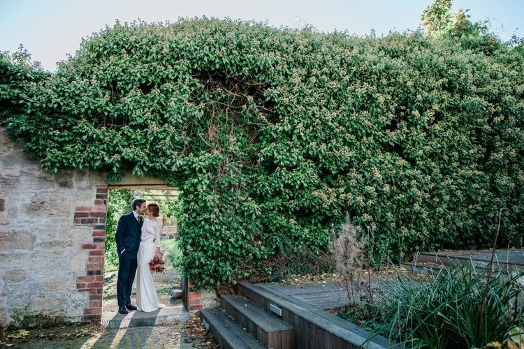 A romantic outdoor wedding portrait captured by Mirrorbox Photography, showing a couple standing together beneath a lush, ivy-covered archway in a peaceful garden setting. The stone walls and greenery create a secluded, intimate frame around the pair