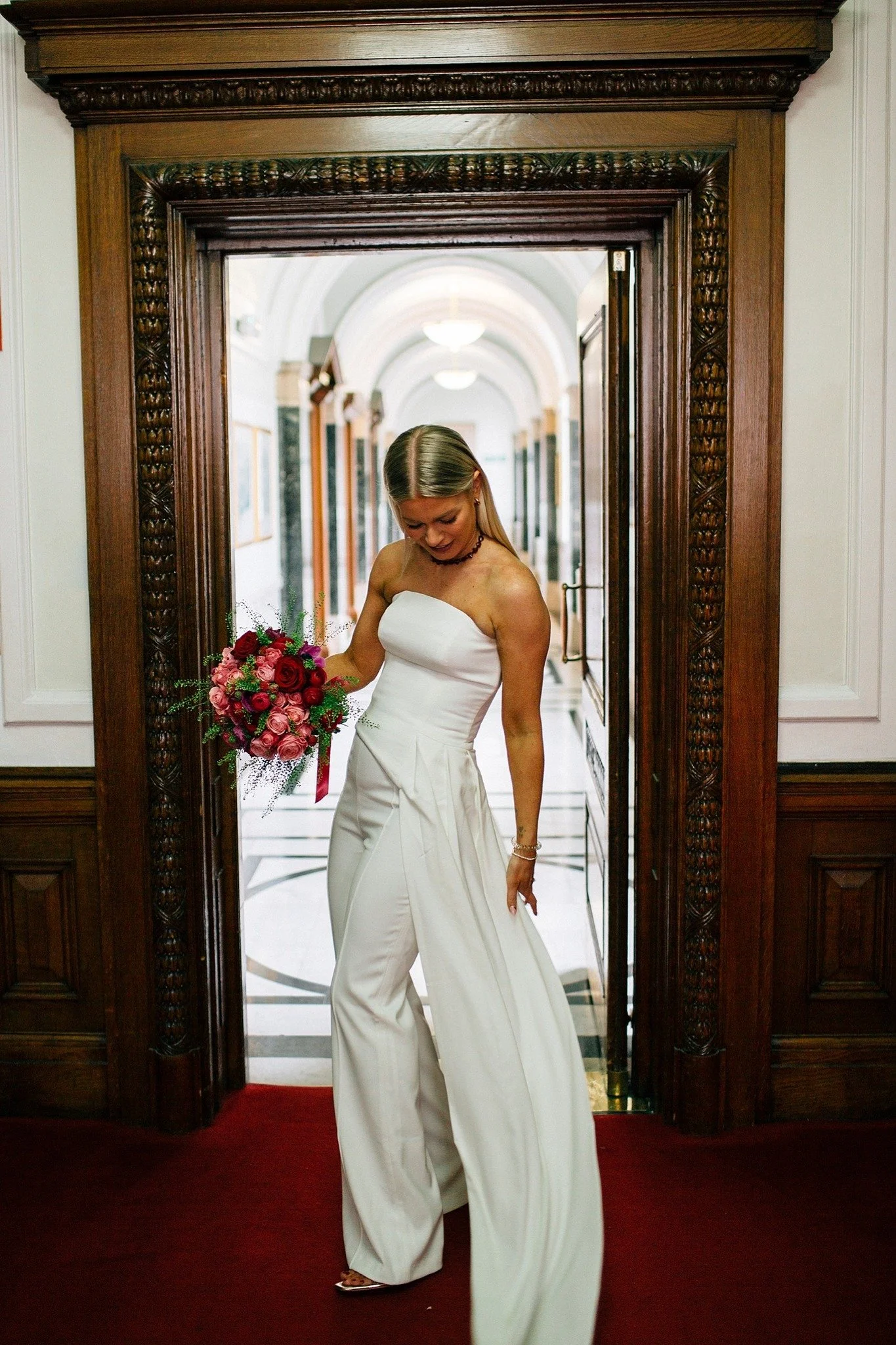 In this image, a bride stands in a doorway framed by dark carved wood. She’s wearing a white, strapless jumpsuit with wide legs and a long overlay panel. She’s holding a bouquet of red and pink roses with greenery, looking down toward the fabric of h