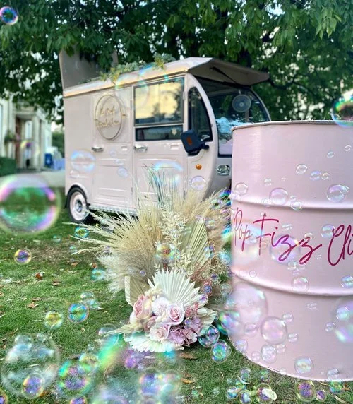 A stylish pale pink prosecco and cocktail van setup featuring a pastel bar drum with “Pop Fizz Clink” lettering, surrounded by soft pampas grass, dried palms, and blush roses. Captured outdoors beneath lush trees, this elegant Pink Drinks Van display