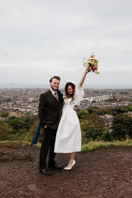A fun, celebratory wedding portrait captured by Mirrorbox Photography, showing a couple standing on a hilltop with sweeping views over the city below. The bride, wearing a short vintage-inspired dress and veil, lifts her bouquet triumphantly into the