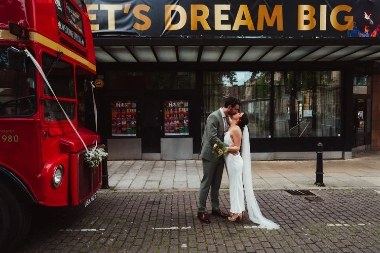 A newly married couple share a romantic kiss outside a theatre with a bold “Let’s Dream Big” sign overhead, creating a cinematic and inspirational backdrop. The bride’s sleek, modern gown and long veil flow elegantly as she stands with her partner, w