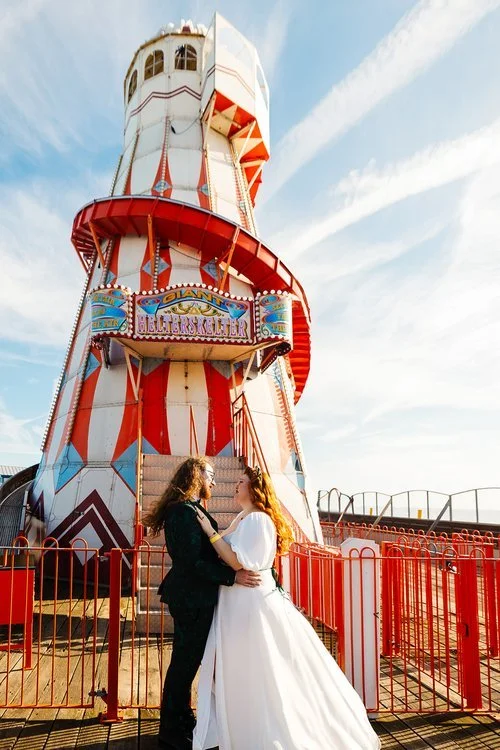 A couple embrace beneath the vibrant red and white structure of a traditional helter skelter, bathed in golden seaside light. The bride’s flowing dress catches the breeze as they look at one another, framed by funfair colours, bold signage, and the o