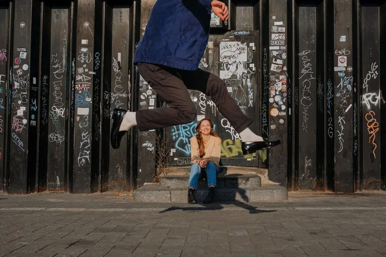 A couple share a fun playful moment in an urban setting as one person jumps mid-air in front of a graffitied backdrop while the other sits smiling on the steps, captured by Joshua Humphrey Photography. The creative framing and street-style atmosphere