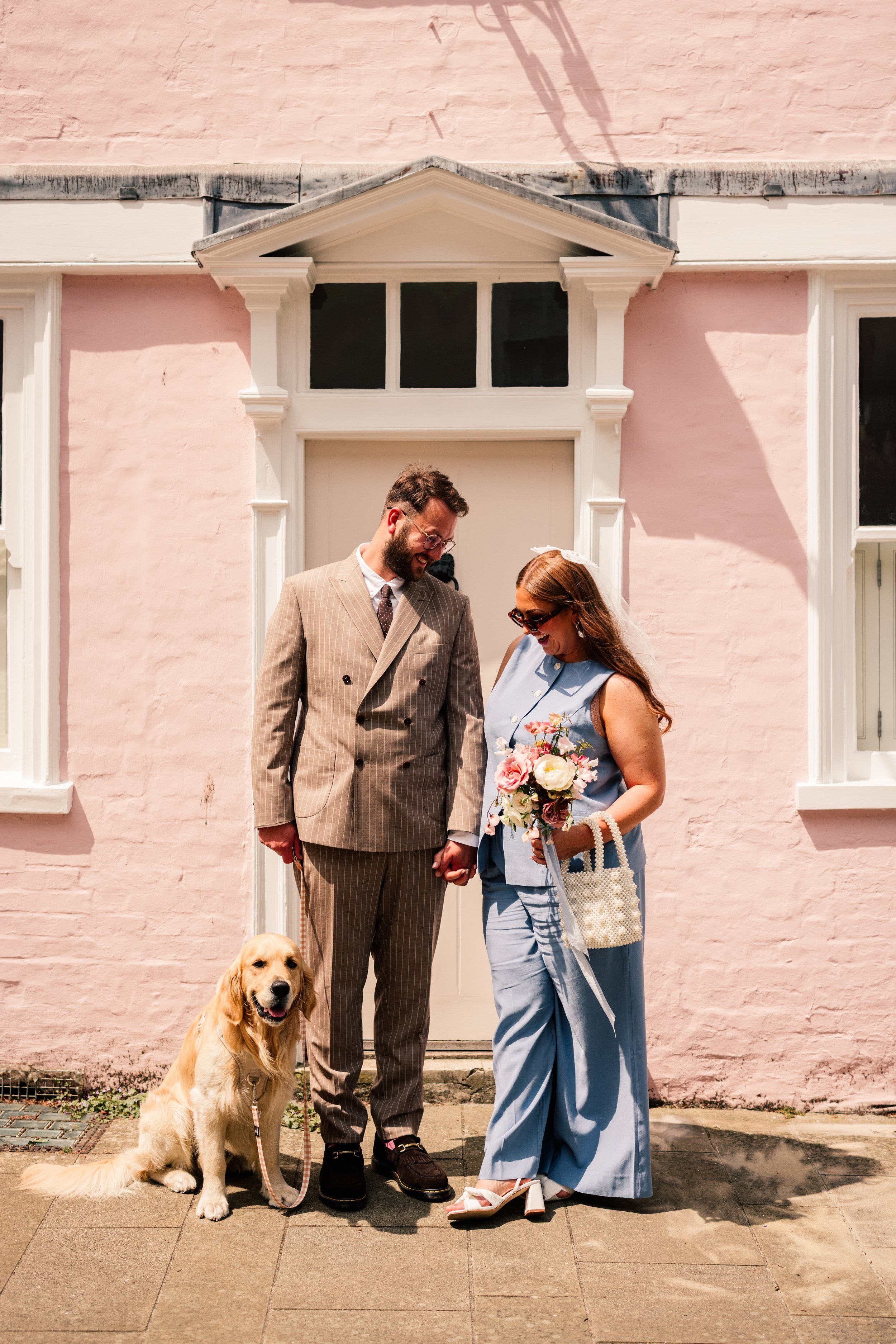 A stylish couple and their golden retriever pose outside a pastel-pink townhouse in Winchester during their modern micro-wedding. The pair hold hands, smiling warmly at each other, with the bride wearing a chic light-blue two-piece suit and veil, and