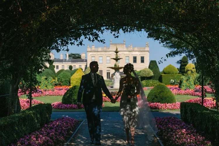 A bride and groom walk hand in hand beneath a shaded garden archway, with vibrant flowerbeds and an elegant historic estate in the background, beautifully captured by Christy Photography. The mix of soft silhouettes, structured topiary and colourful 