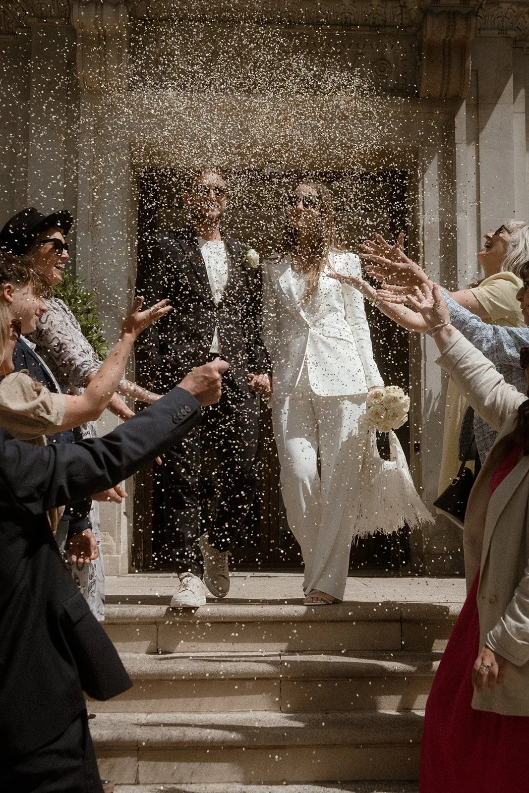 A newly married couple walks down stone steps outside a city hall while guests throw a large amount of confetti that fills the air, with the bride in a tailored white suit holding a bouquet and the groom in a dark suit, surrounded by friends and fami