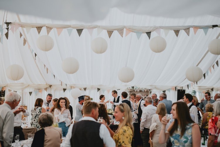 A lively and atmospheric reception scene captured by Matt Fox Photography, showing guests gathered inside a beautifully decorated marquee. Soft white draping, pastel bunting and hanging paper lanterns create a relaxed and elegant setting as friends a
