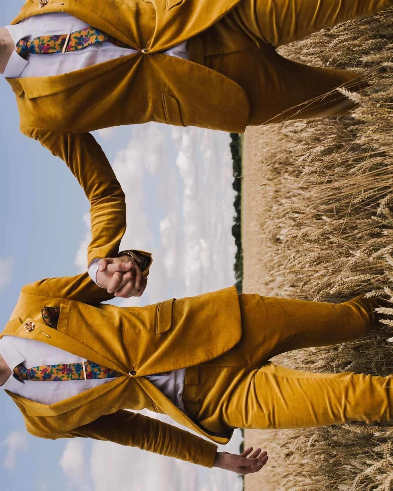 An artistic photo of 2 grooms on their wedding day walking hand in hand through a corn field. They are wearing mustard, velvet suits and floral ties. There is blue sky in the background, and the image is on it's side.