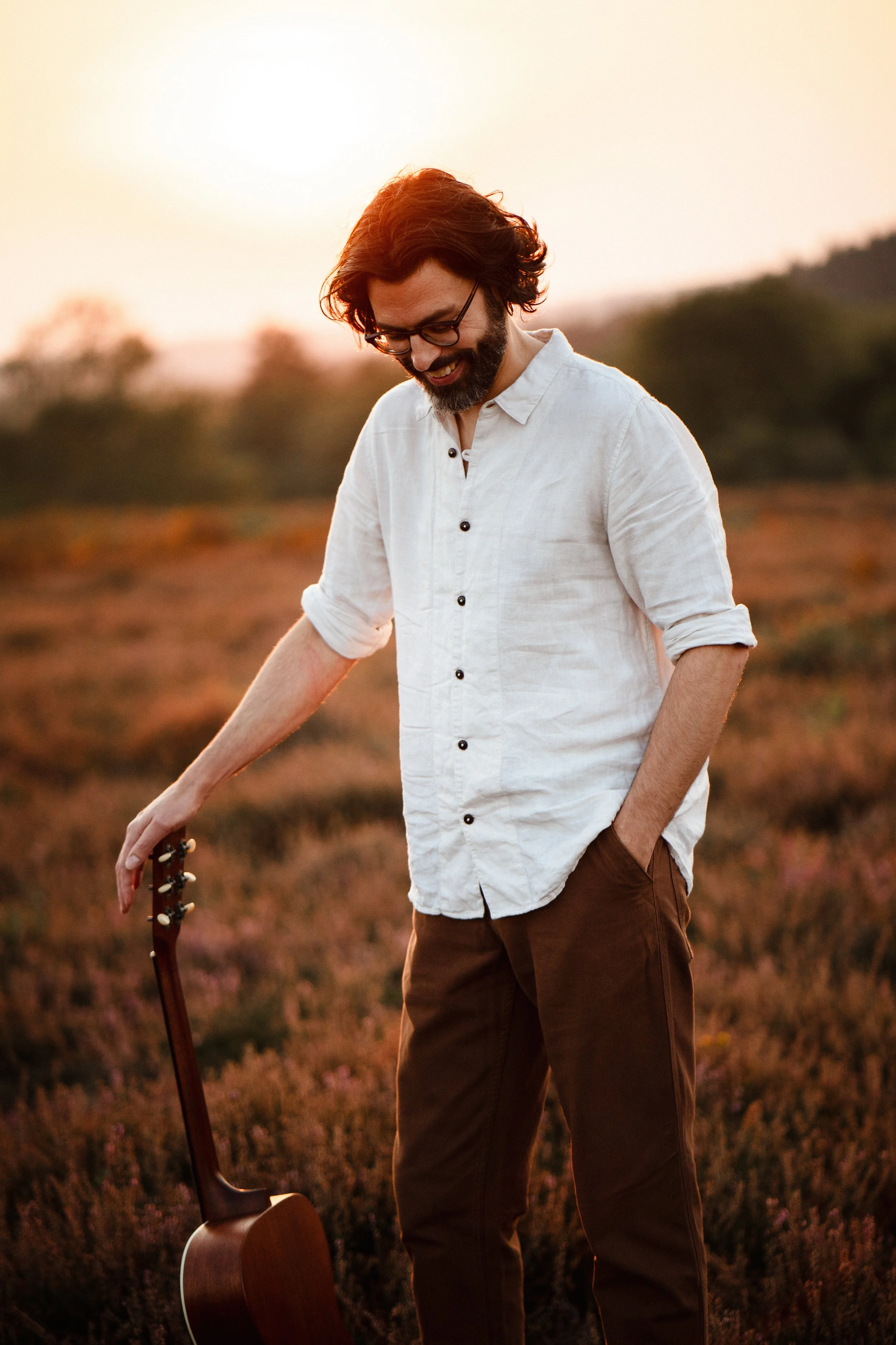 Thomas J Wilman, an acoustic wedding singer and guitarist, stands in a field at sunset holding his guitar. His relaxed, natural style captures the warmth and authenticity of his live acoustic wedding performances. Clarkie Photography.