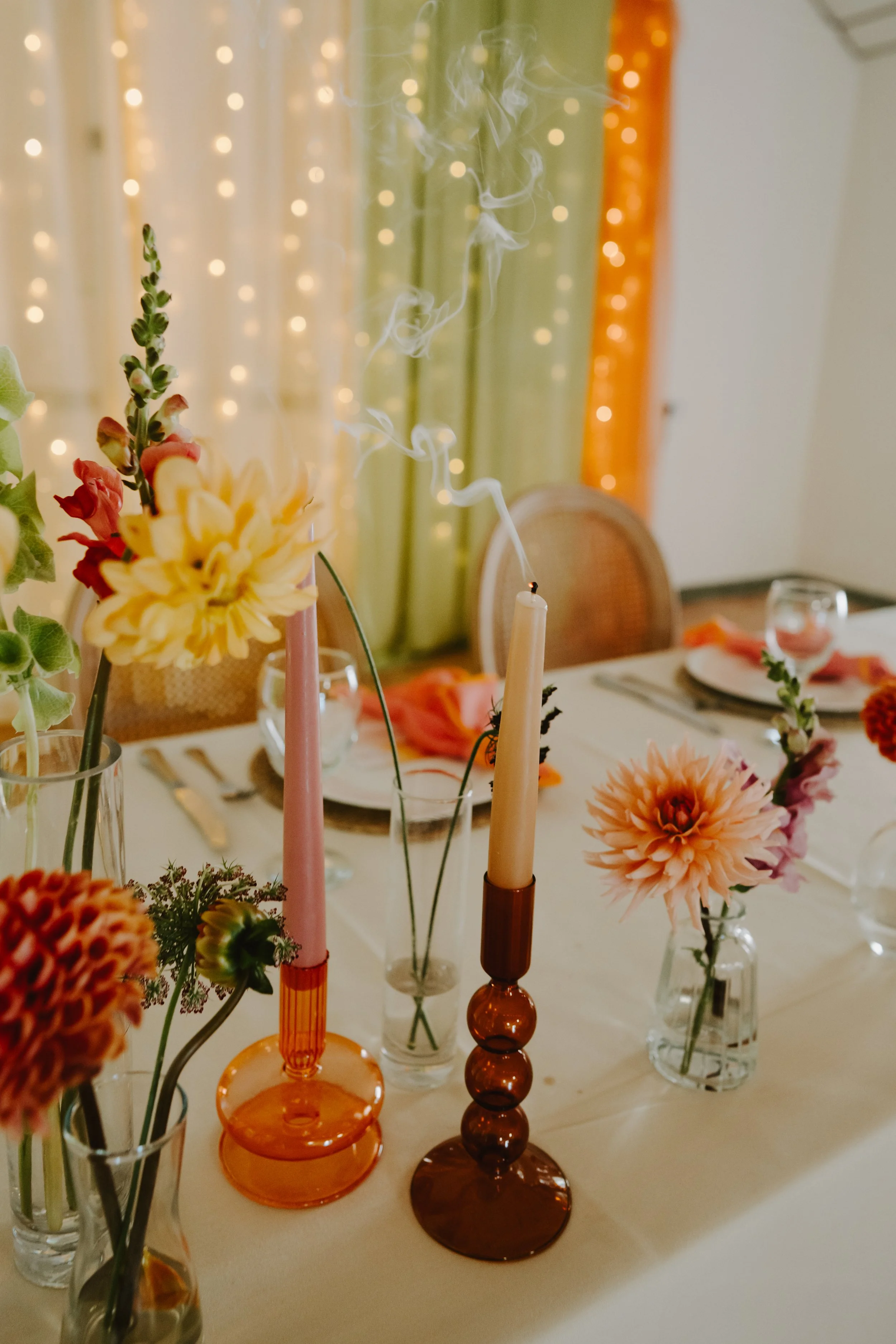 Orange Tree Weddings styling and décor showcasing a colourful wedding table with sculptural glass candlesticks, pastel taper candles, seasonal florals and soft fairy-light backdrops for a warm, modern celebration atmosphere.