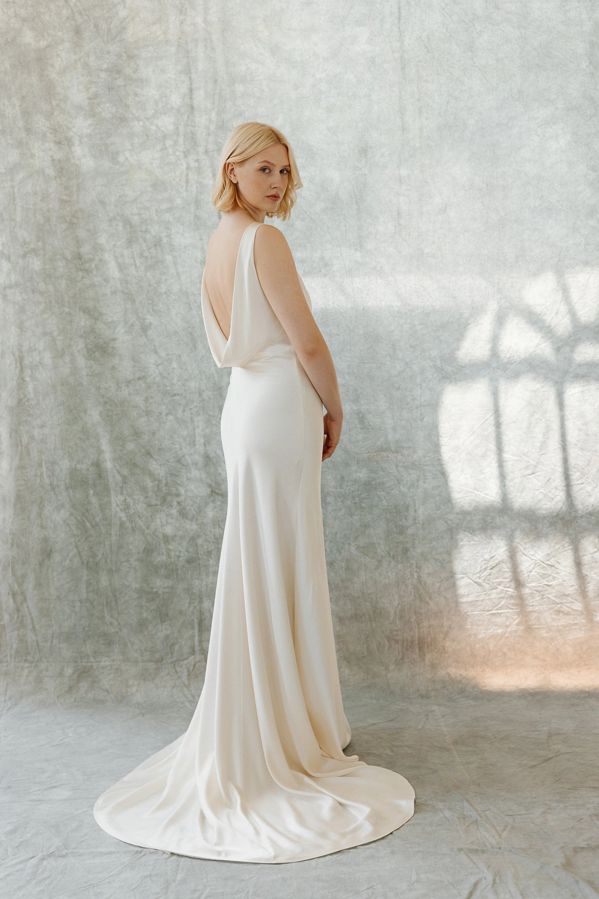 The bride stands against a softly textured backdrop wearing a sleek bias cut silk gown with a long fluid train, the low draped cowl back creating an elegant silhouette as gentle light falls across the scene and highlights the smooth simplicity of the