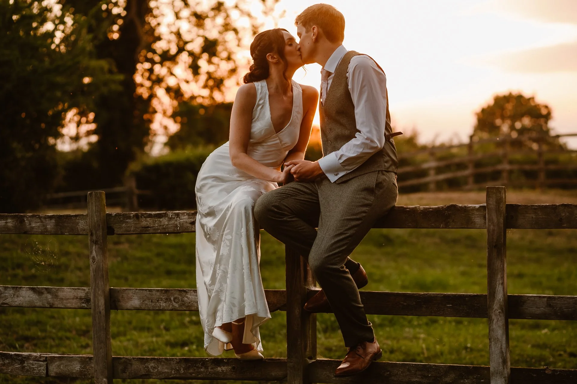 A romantic countryside wedding photo captured by Bluebell Photo Studio, showing a bride and groom sharing an intimate kiss at golden hour in a lush green field surrounded by a flock of sheep. The groom dips the bride as her embroidered gown and long 