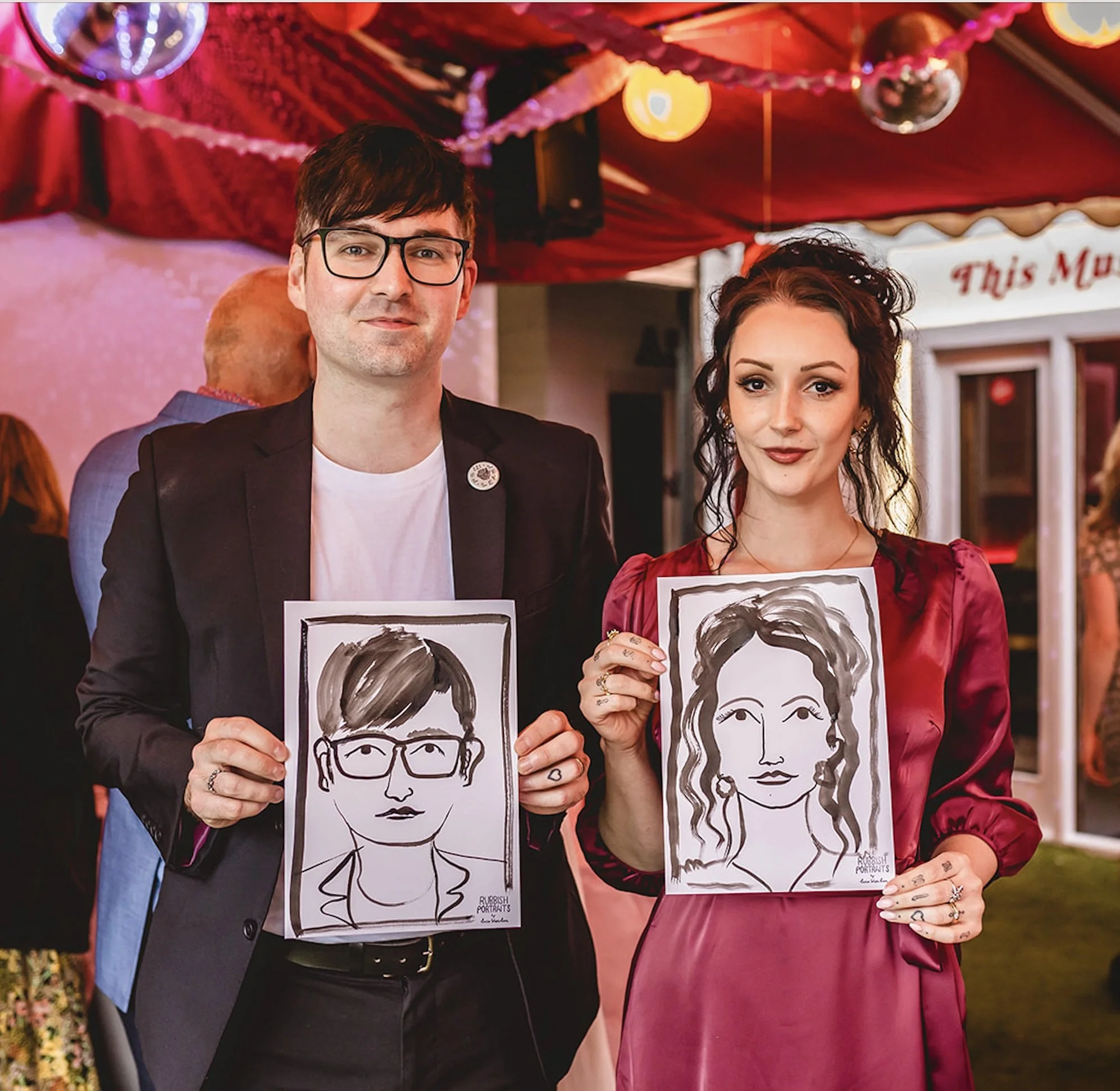 Event guests holding hand-drawn portraits from the Rubbish Portraits photo booth at a stylish celebration under a red marquee. The pair pose proudly with their custom black and white caricatures. Perfect inspiration for wedding entertainment, live ev