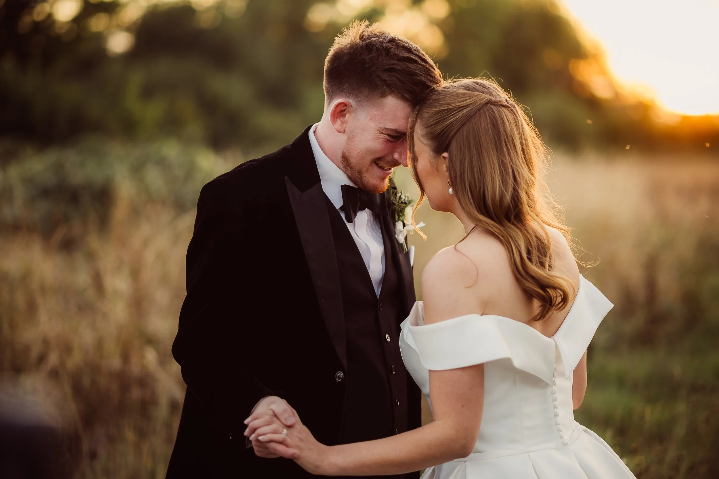 A couple shares an intimate moment in the golden light of sunset, their foreheads gently touching as they hold hands. The groom wears a black tuxedo with a velvet jacket while the bride glows in an elegant off the shoulder gown with soft curls fallin
