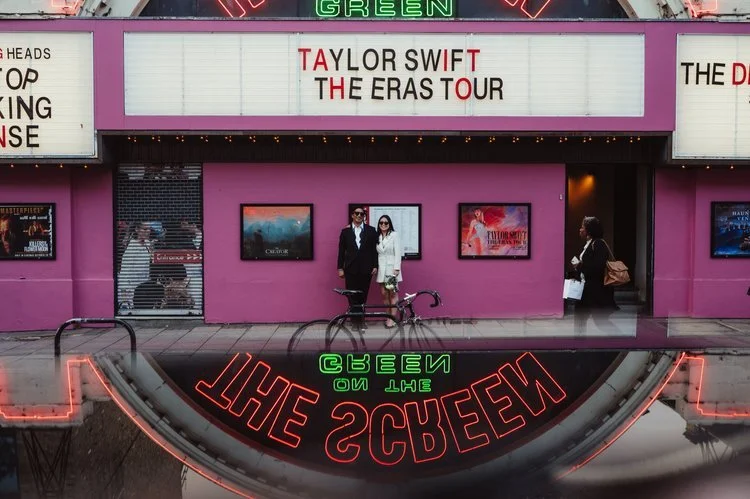 A stylish couple stand outside a vibrant pink cinema façade featuring bold marquee signage and neon reflections, creating a striking and modern urban backdrop. The pair pose confidently beneath the illuminated lettering, surrounded by film posters an