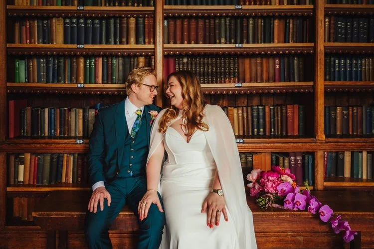 A newly married couple sit together in an elegant library, surrounded by tall wooden bookshelves filled with colourful vintage books. The bride, wearing a modern white gown with a flowing cape, smiles warmly at her partner as they lean in close, crea