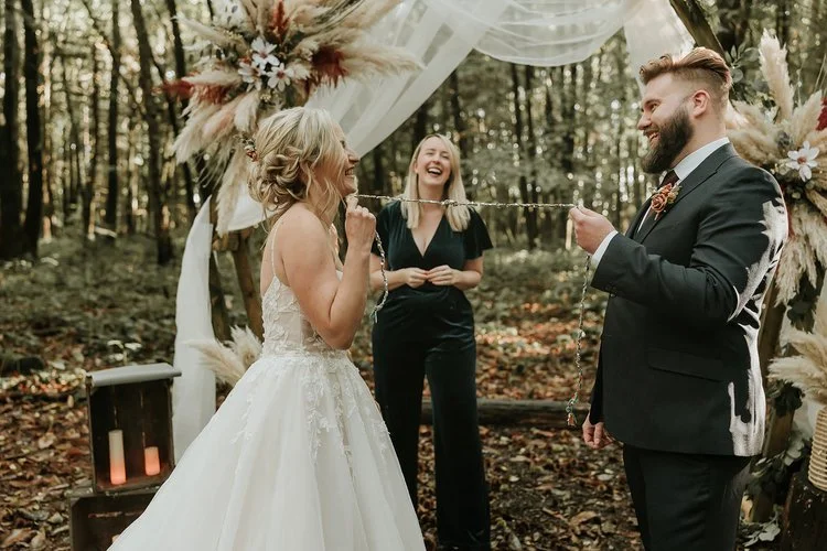 A fun and heartfelt woodland wedding ceremony with a couple laughing as they take part in a hand-fasting ritual, led by their wedding celebrant. The bride wears a flowing white gown, the groom a dark suit with autumnal tones, and wedding celebrant Fr