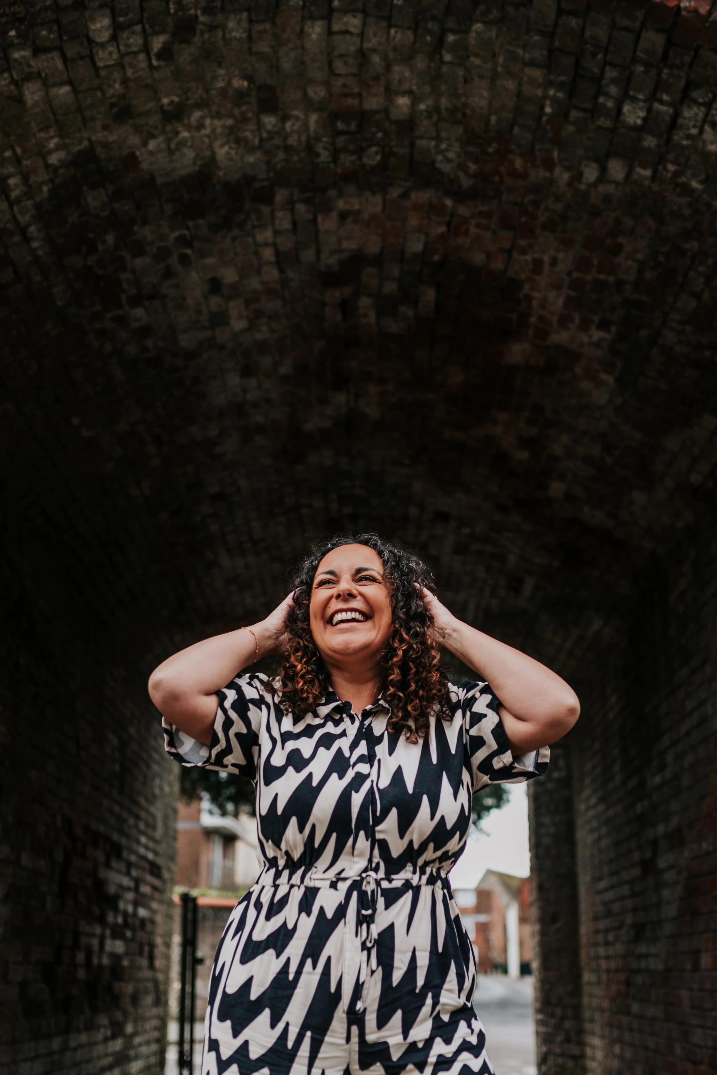 A joyful portrait of a woman laughing beneath a rustic brick archway, her hands lifted to her head in pure delight. Wearing a bold black-and-white patterned jumpsuit, she stands confidently against the textured tunnel backdrop, the soft natural light