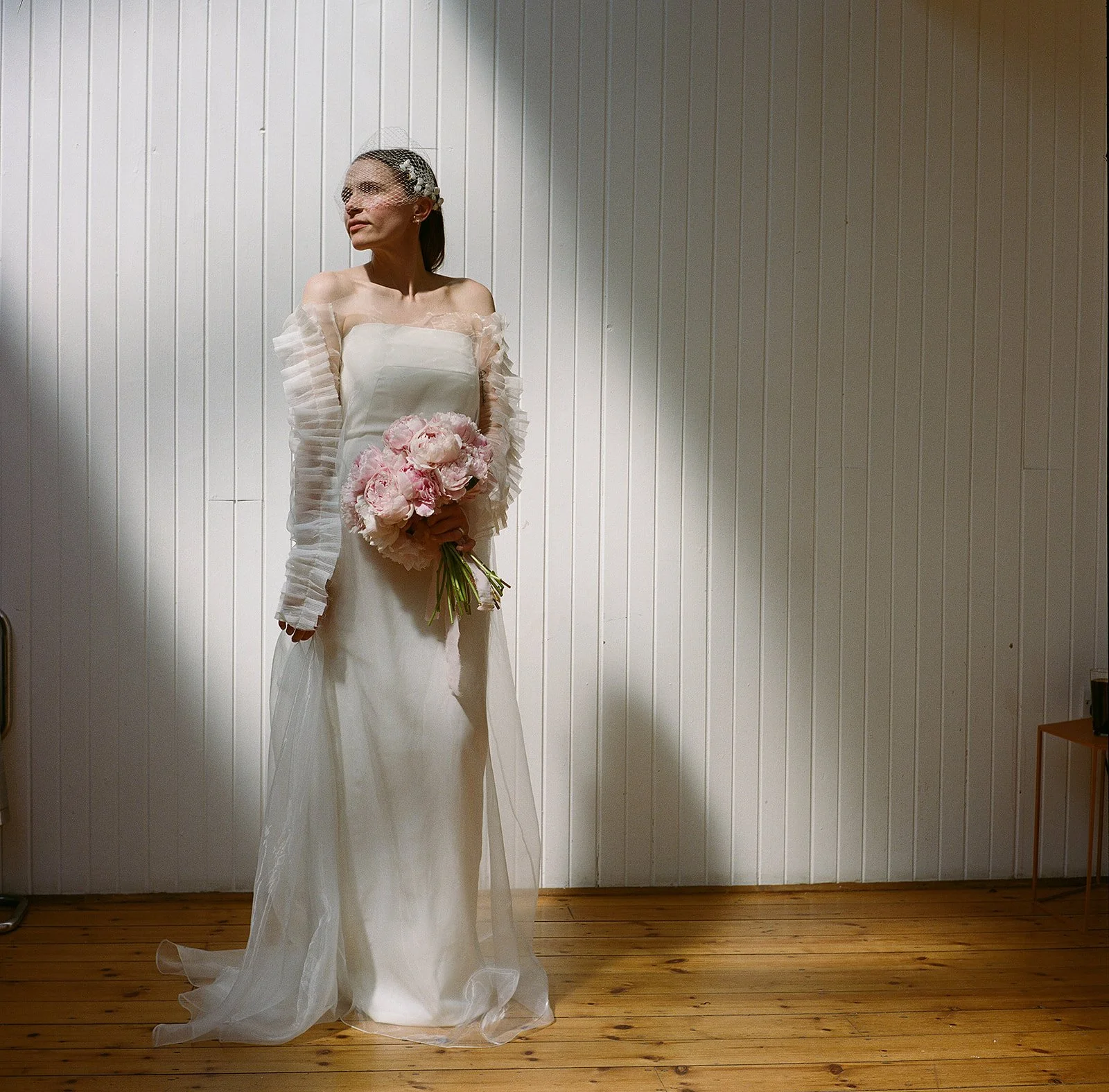 A person stands against a white, vertically panelled wall, wearing a strapless white dress with a straight neckline and sheer, pleated sleeves that fall from the shoulders. A soft layer of translucent fabric drapes over the skirt and pools lightly at