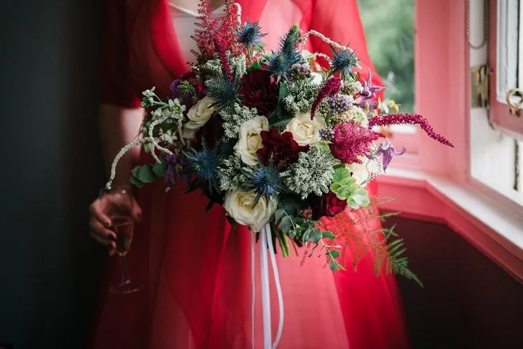Bold romantic wedding bouquet by New Bud Floristry featuring ivory garden roses, deep red blooms, blue thistles and textured seasonal foliage with accents of amaranthus. Held by the bride in a striking red dress beside a softly lit window, this drama