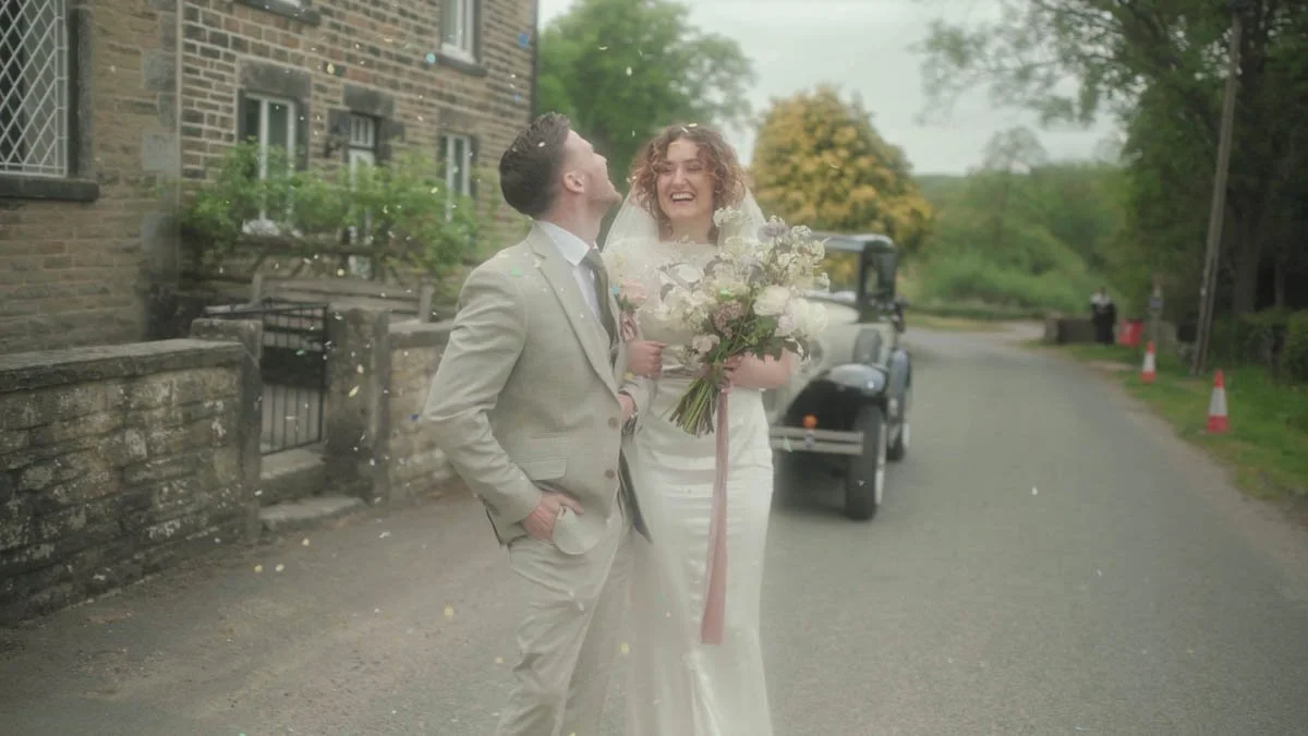 A couple stands laughing on a quiet country road as soft confetti drifts around them and an old-fashioned car waits behind creating a gentle cinematic moment filled with joy tender movement and the relaxed charm of an English village backdrop.