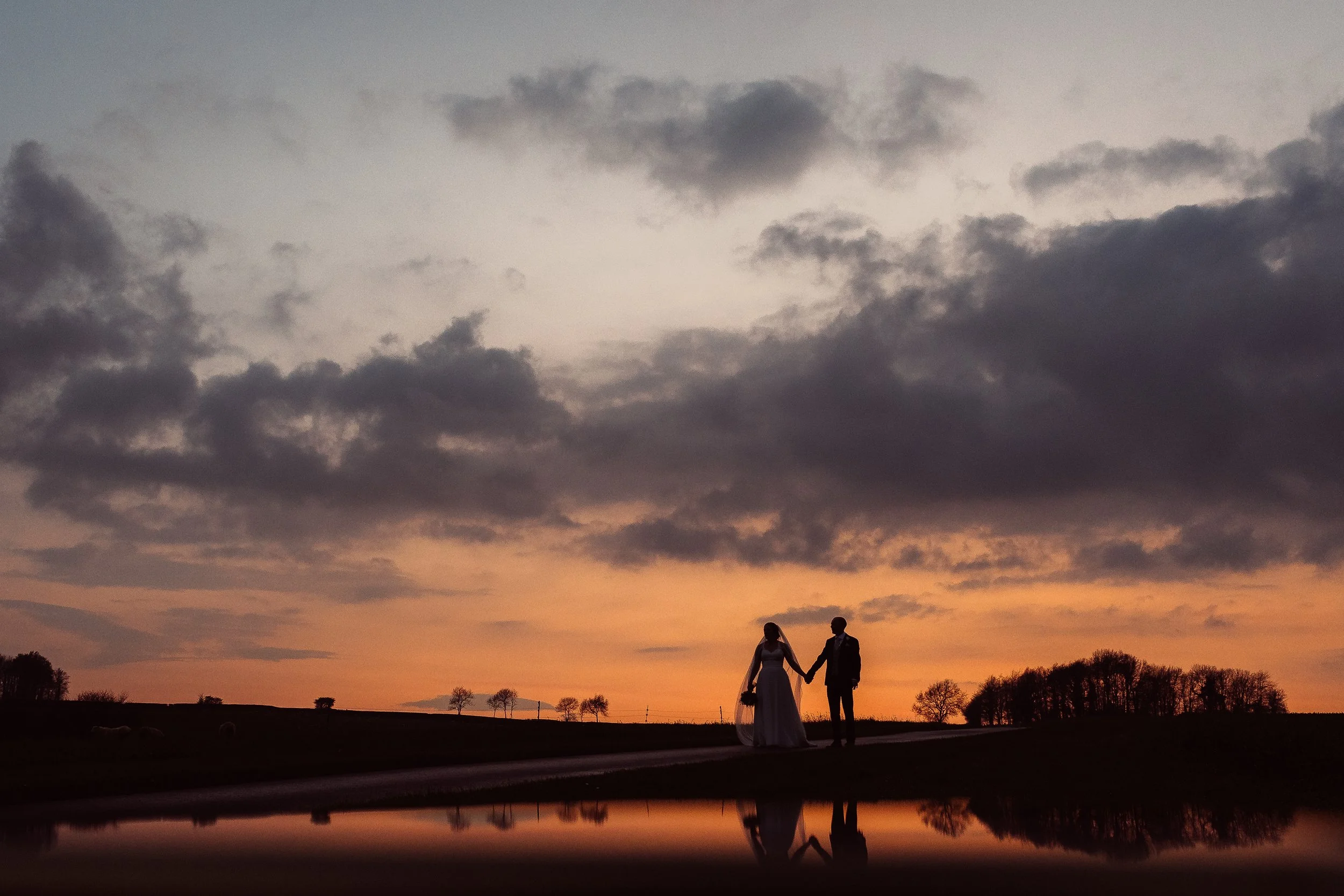 A dramatic sunset wedding silhouette captured by Gemma Gaskins Photography, showing a couple holding hands against a glowing orange sky. Their reflection in the still water adds a cinematic, romantic feel to this artistic outdoor portrait, perfect fo
