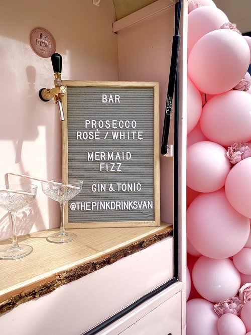 A close-up of The Pink Drinks Van bar setup, featuring a pastel letter board menu listing prosecco, rosé, white wine, gin and tonic, and the signature Mermaid Fizz. Two coupe glasses sit on the wooden counter, with soft pink balloons in the backgroun