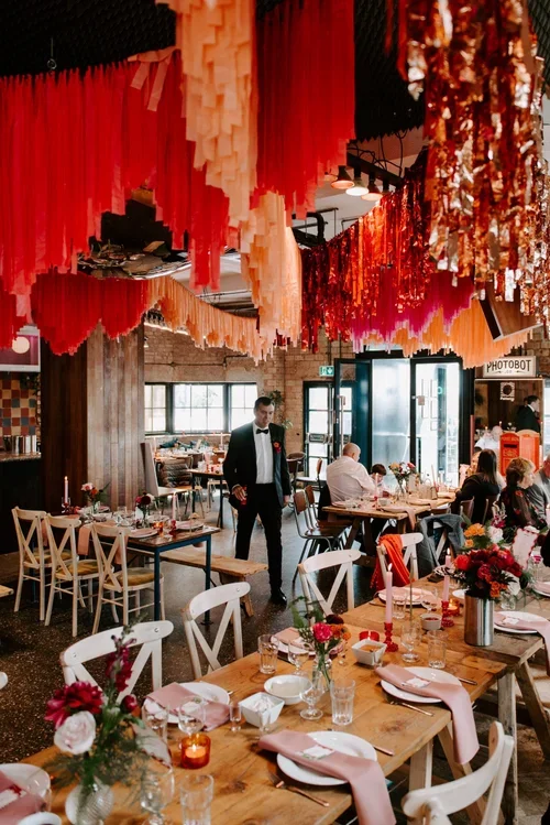 A vibrant wedding reception setup at Patterns Brighton, featuring bold red, orange, and pink ceiling streamers that add a playful retro vibe to the modern industrial venue. Rustic wooden tables are styled with pink napkins, colorful floral arrangemen