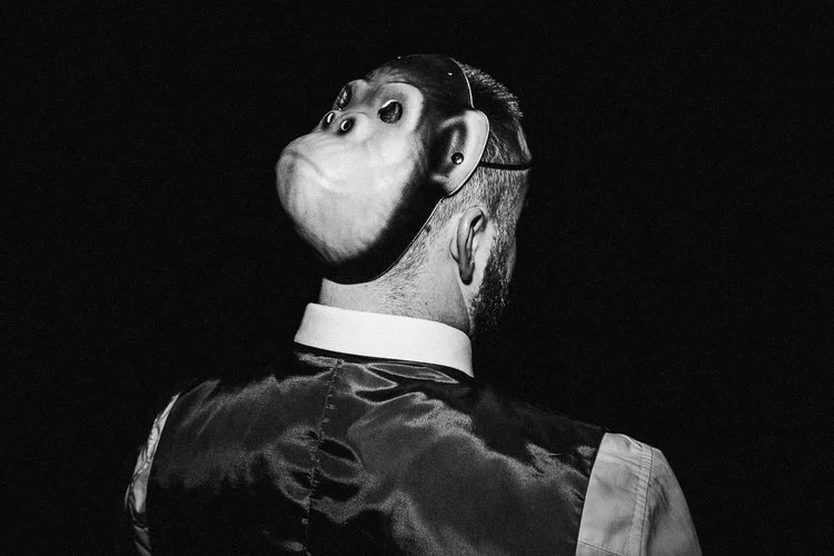 A dramatic black and white photograph by Matt Parry Photography that captures the back of a man wearing a playful monkey mask during what appears to be a lively wedding celebration. The subject is dressed in a formal outfit with a crisp white shirt a