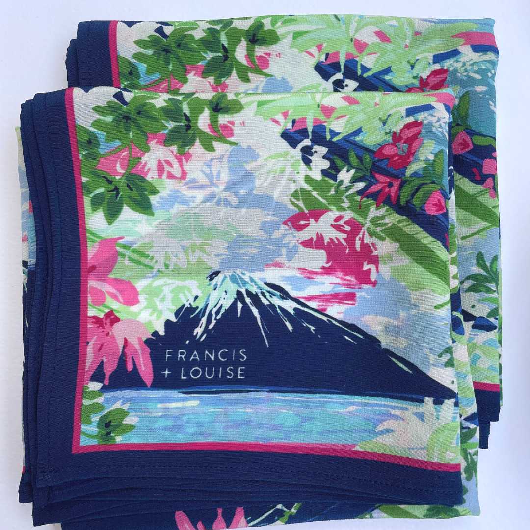 A neatly folded Francis + Louise pocket square featuring a vibrant illustrated design with lush green leaves, pink flowers and a stylised dark blue mountain rising above a light blue body of water. The pattern is framed by a deep blue border with a t