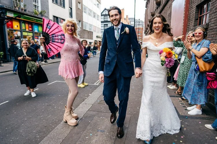 A newly married couple walk hand in hand through a lively city street, surrounded by cheering onlookers and vibrant characters, beautifully captured by Jordanna Marston Photography. The bride’s bright bouquet and joyful expression, paired with the co