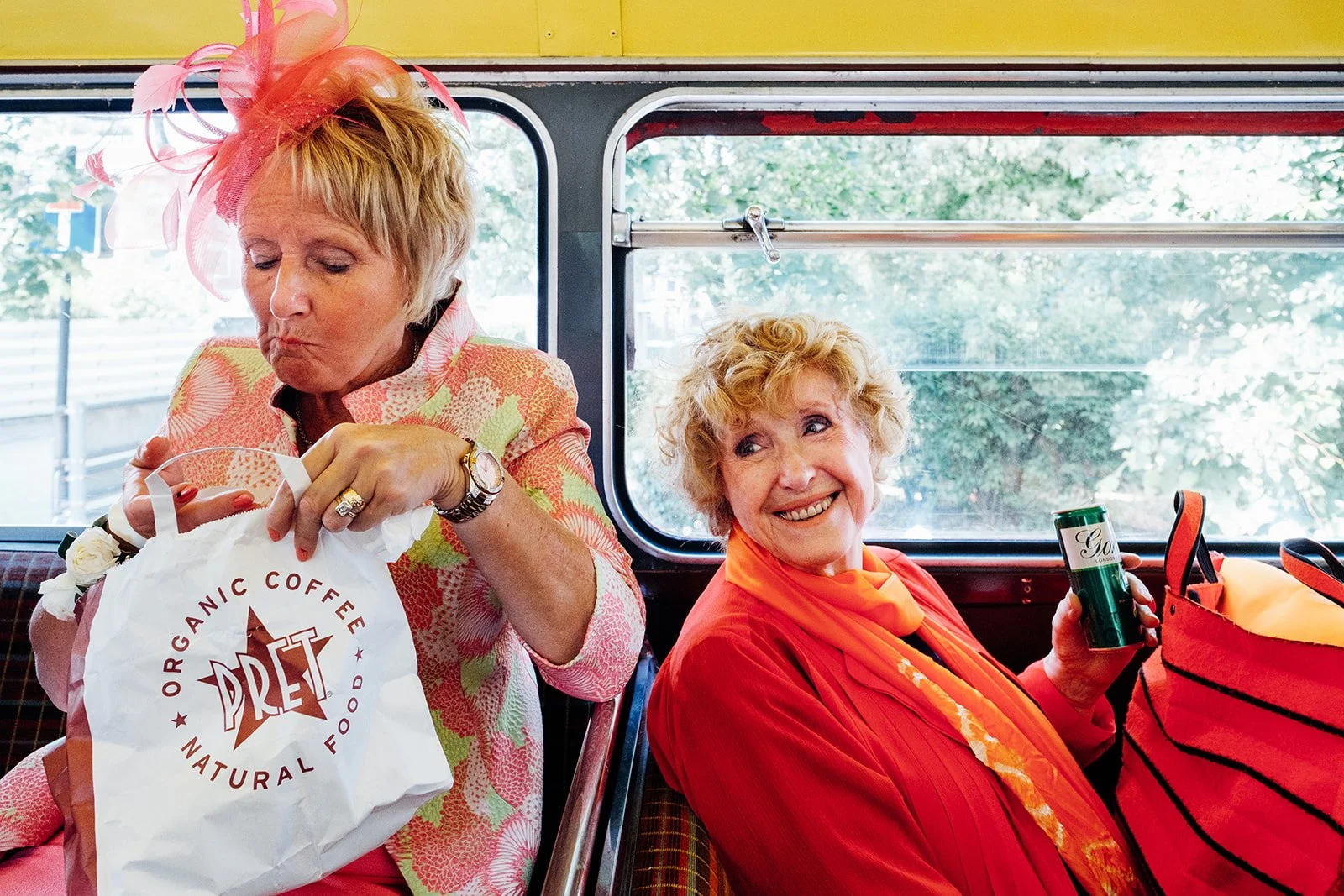 Two women dressed in bright formal outfits are seated on a bus, with one woman opening a Pret bag while wearing a decorative coral fascinator and the other smiling warmly while holding a canned drink beside an orange striped bag. Sunlight filters thr