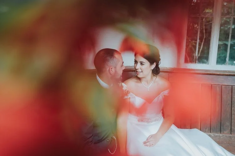 A romantic and softly artistic wedding portrait captured by Matt Fox Photography, showing a couple sharing an intimate moment while framed by vibrant out of focus foliage. The bride, dressed in an elegant off the shoulder gown with delicate jewellery