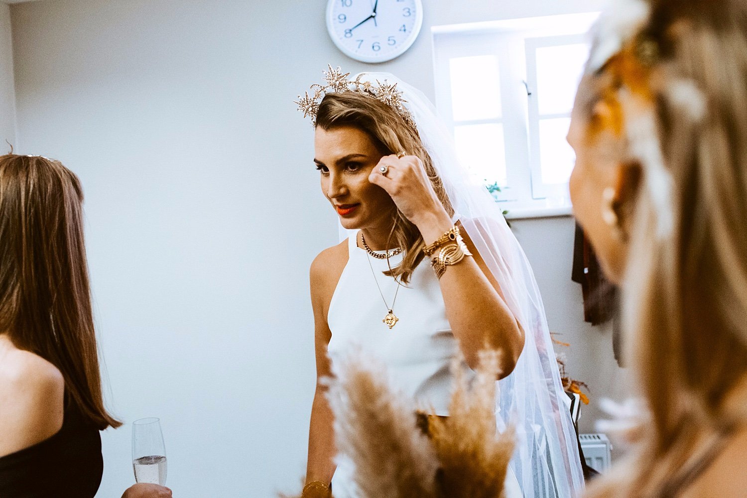 A candid bridal prep moment captured by Paola de Paola Photography shows a stylish bride in a modern high-neck white wedding dress adjusting her celestial gold crown while chatting with friends. Her bold red lipstick, layered gold jewellery and relax