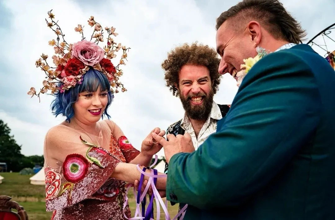 A joyful, festival-style wedding ceremony led by celebrant Lonan Jenkins, capturing colourful outfits, live music, laughter and heartfelt moments as guests celebrate love in a relaxed, inclusive outdoor setting.