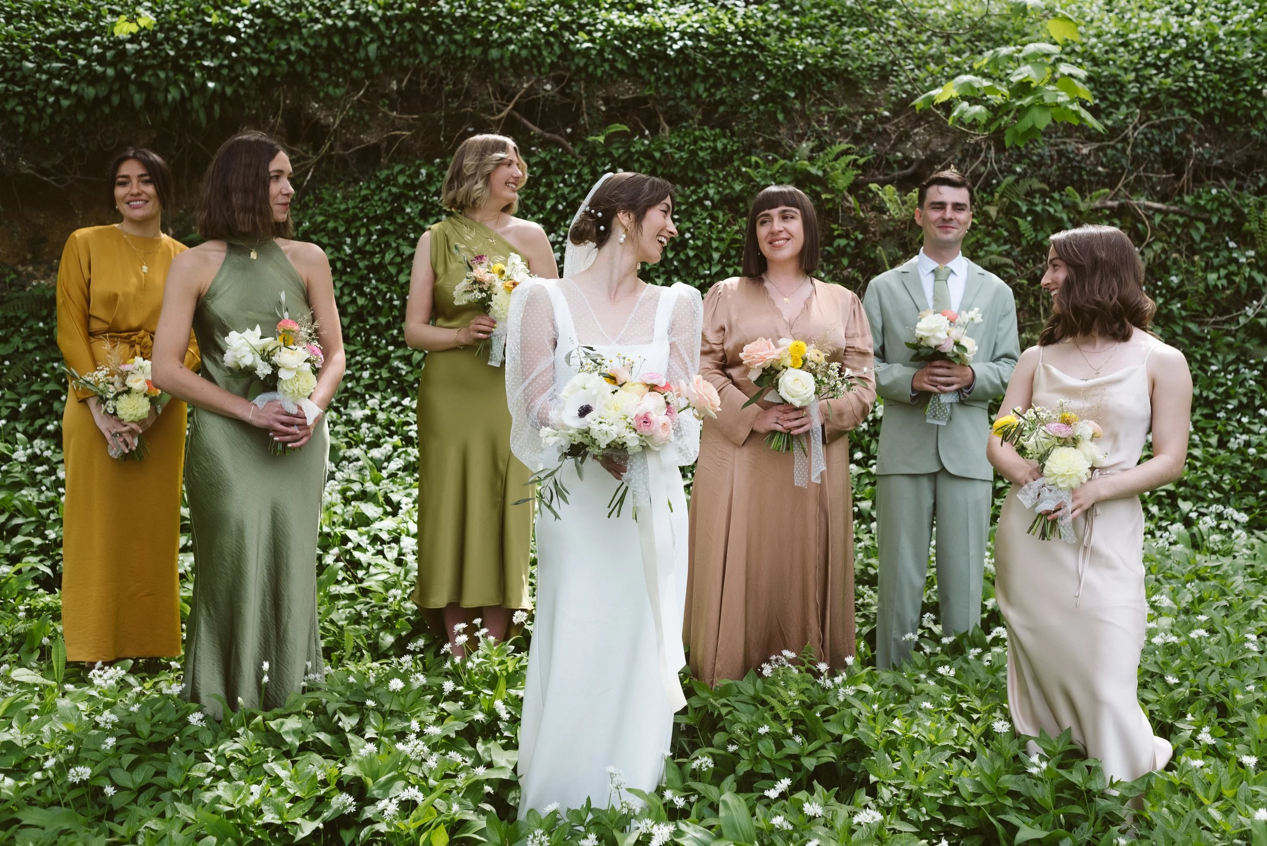 A bride stands surrounded by her wedding party in a lush green woodland setting, captured by Roz Pike Photography. Their coordinated pastel and earth-tone outfits, natural bouquets and relaxed smiles create a stylish contemporary portrait with a soft
