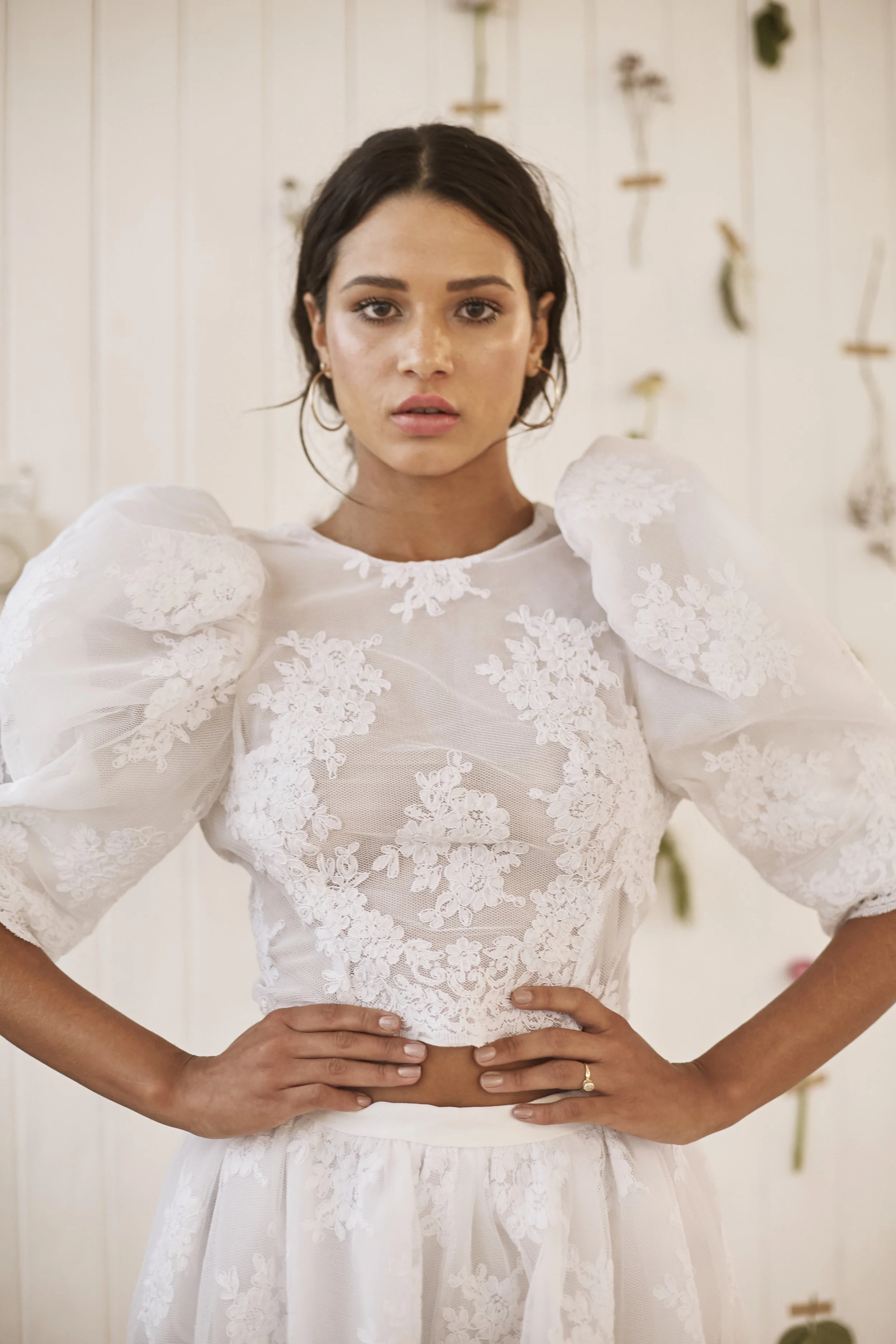 A person faces the camera wearing a fitted white top with sheer fabric and floral lace detailing, featuring puffed sleeves and a high neckline. Their hands rest at the waist, drawing attention to the texture and structure of the garment. The backgrou