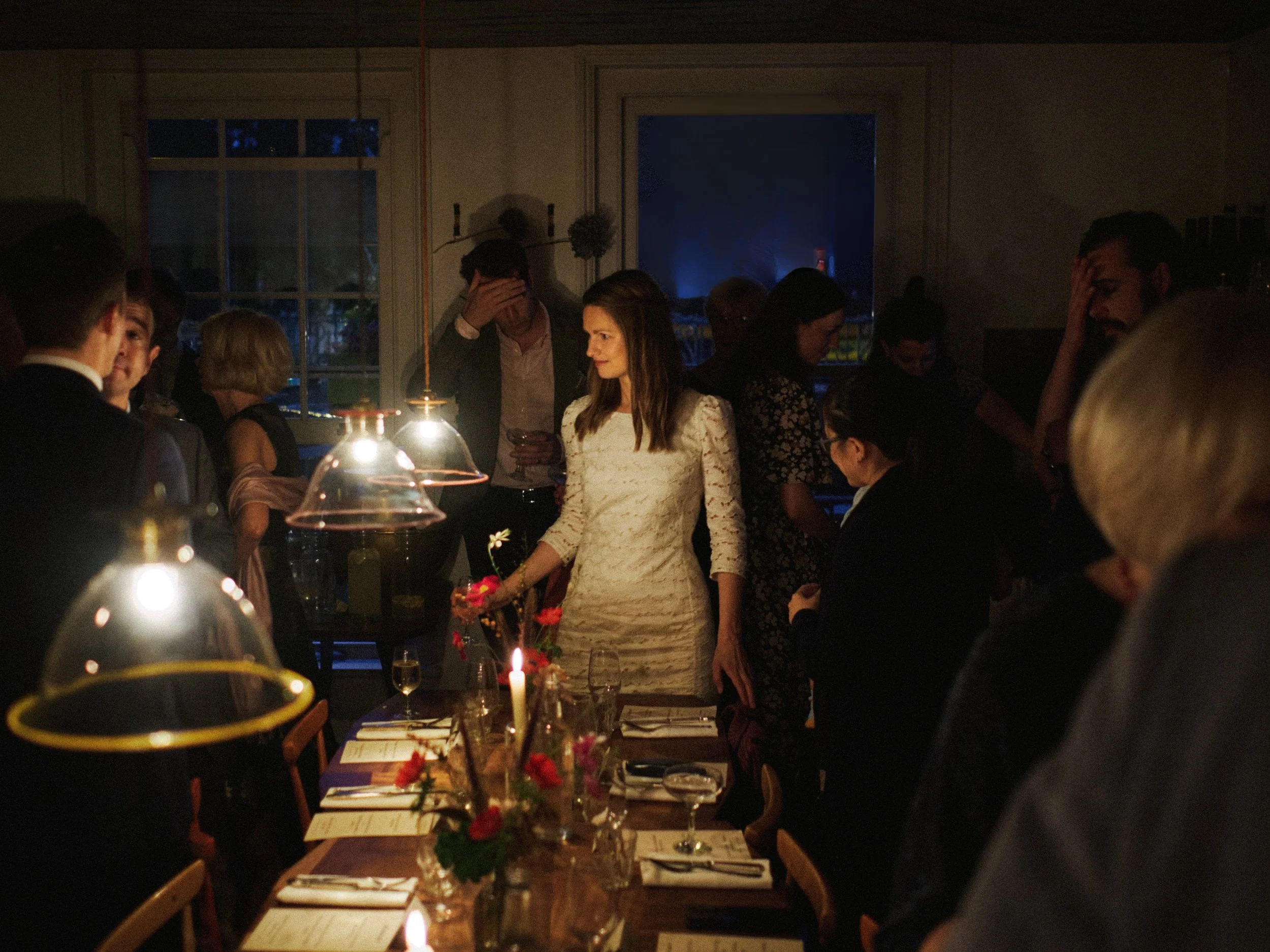 A warm and atmospheric documentary image captured by Wayne La Photography, showing the bride standing at the head of a candlelit dinner table as guests gather around her. Soft pools of golden light from hanging lamps illuminate the scene, creating a 