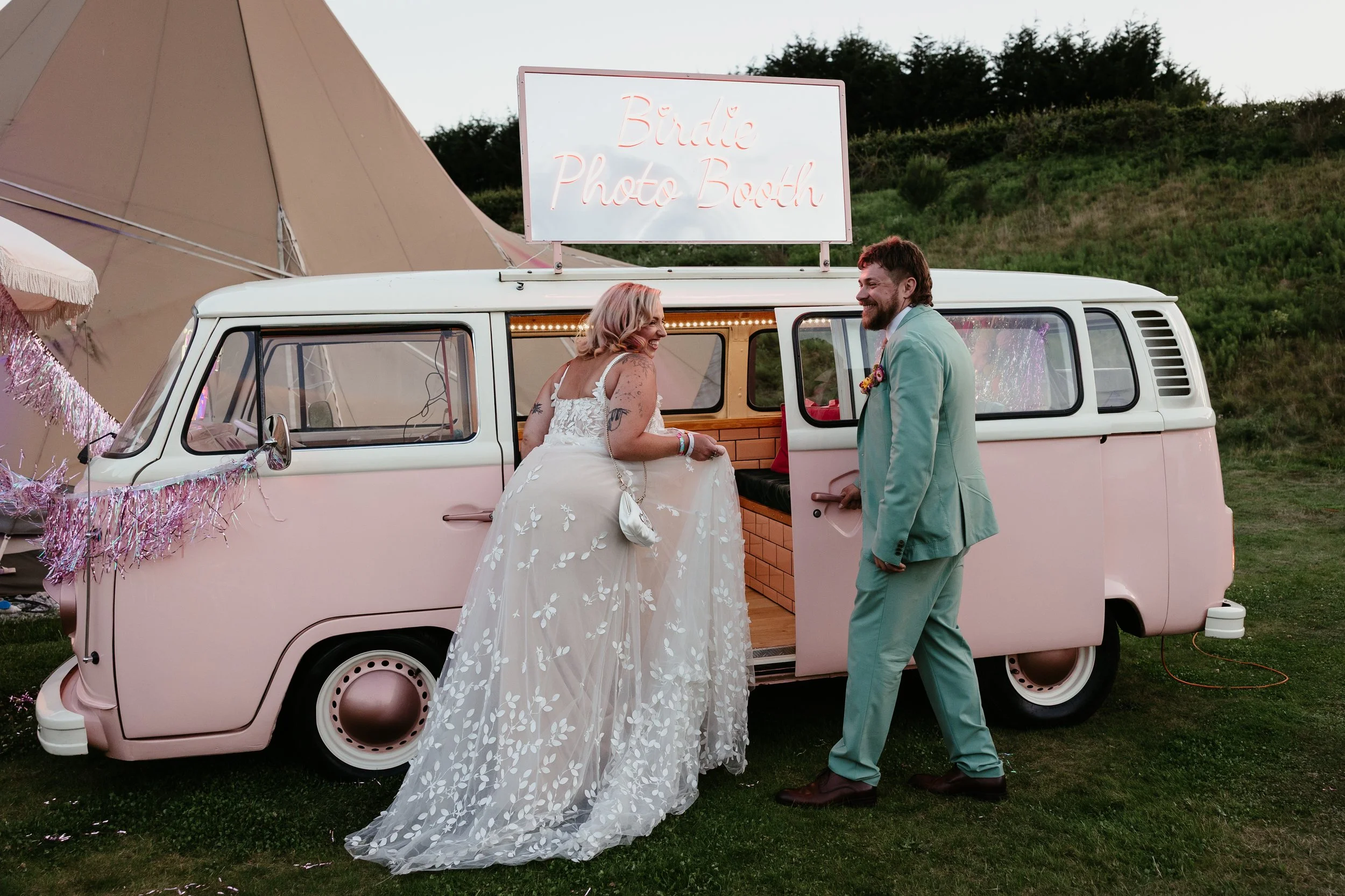 A fun and stylish wedding moment captured by Joy Louise Photography, showing the couple arriving at a retro pink campervan photo booth during their outdoor celebration. The bride wears a beautiful embroidered gown with a flowing train as she steps to