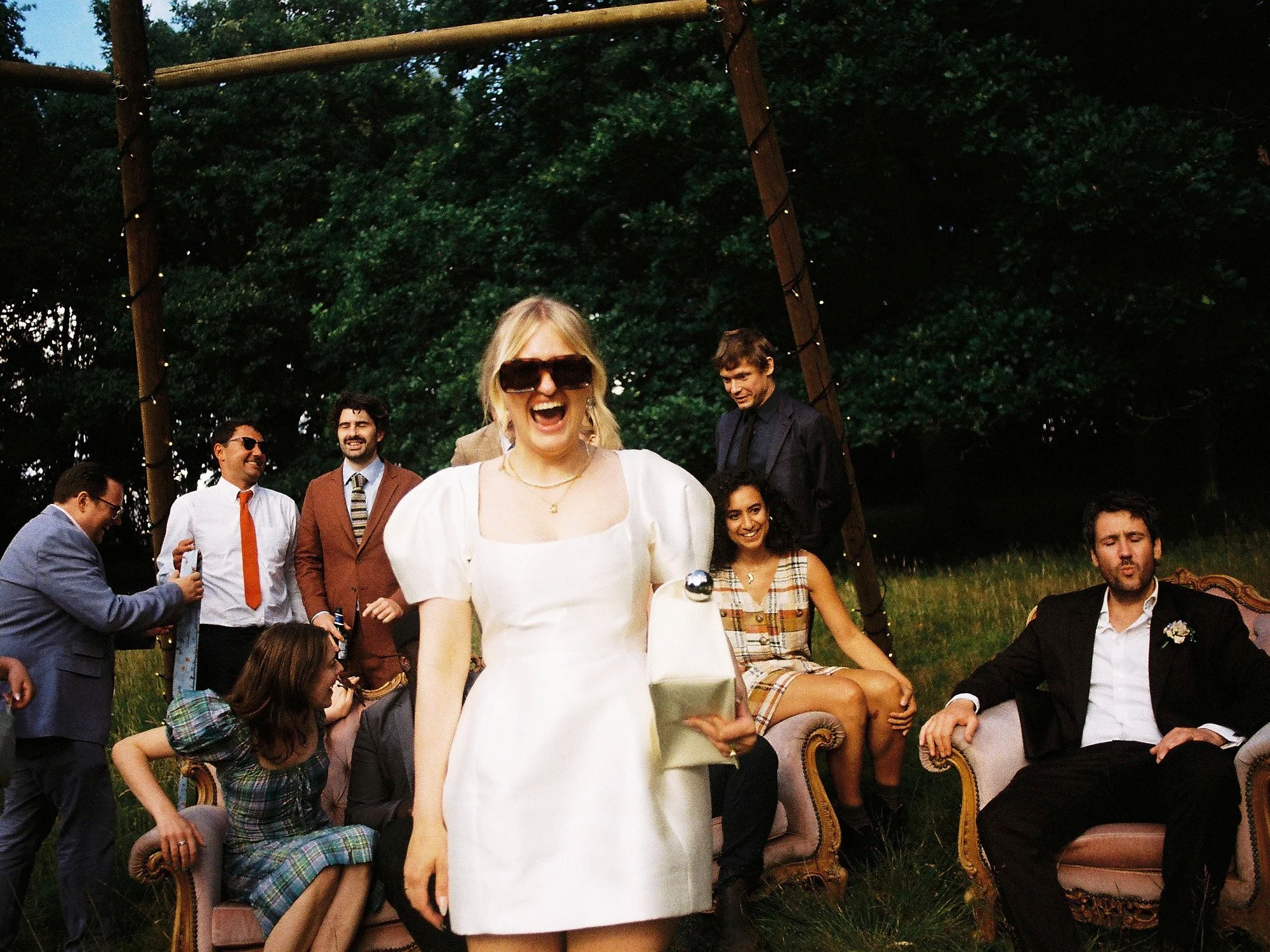 A lively and cinematic candid captured by Wayne La Photography, showing a bride moving confidently toward the camera as a joyful crowd gathers behind her in an outdoor evening setting. Her bold sunglasses and modern white dress create a playful, fash