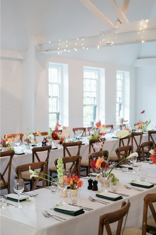 Bright modern wedding tablescape featuring colourful floral arrangements by Botanique Workshop with vibrant seasonal stems displayed in petite vases along long banquet tables. Set within a light filled venue with wooden chairs, crisp white linens and