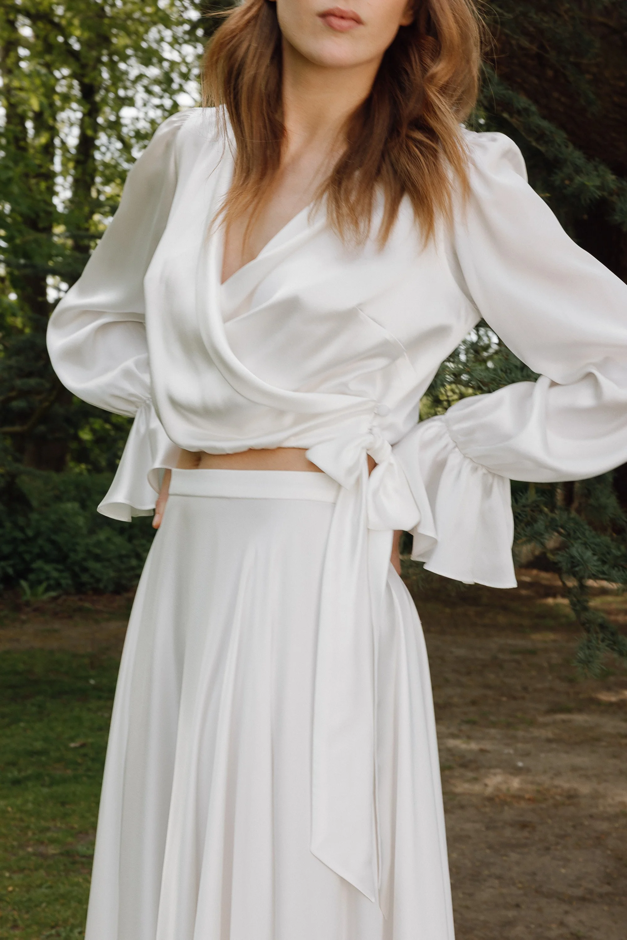 The bride stands outdoors surrounded by soft greenery while wearing flowing silk separates that catch the natural light, the wrap top gathered at the waist with long ties and gently billowed sleeves that mirror the movement of the smooth, full skirt 