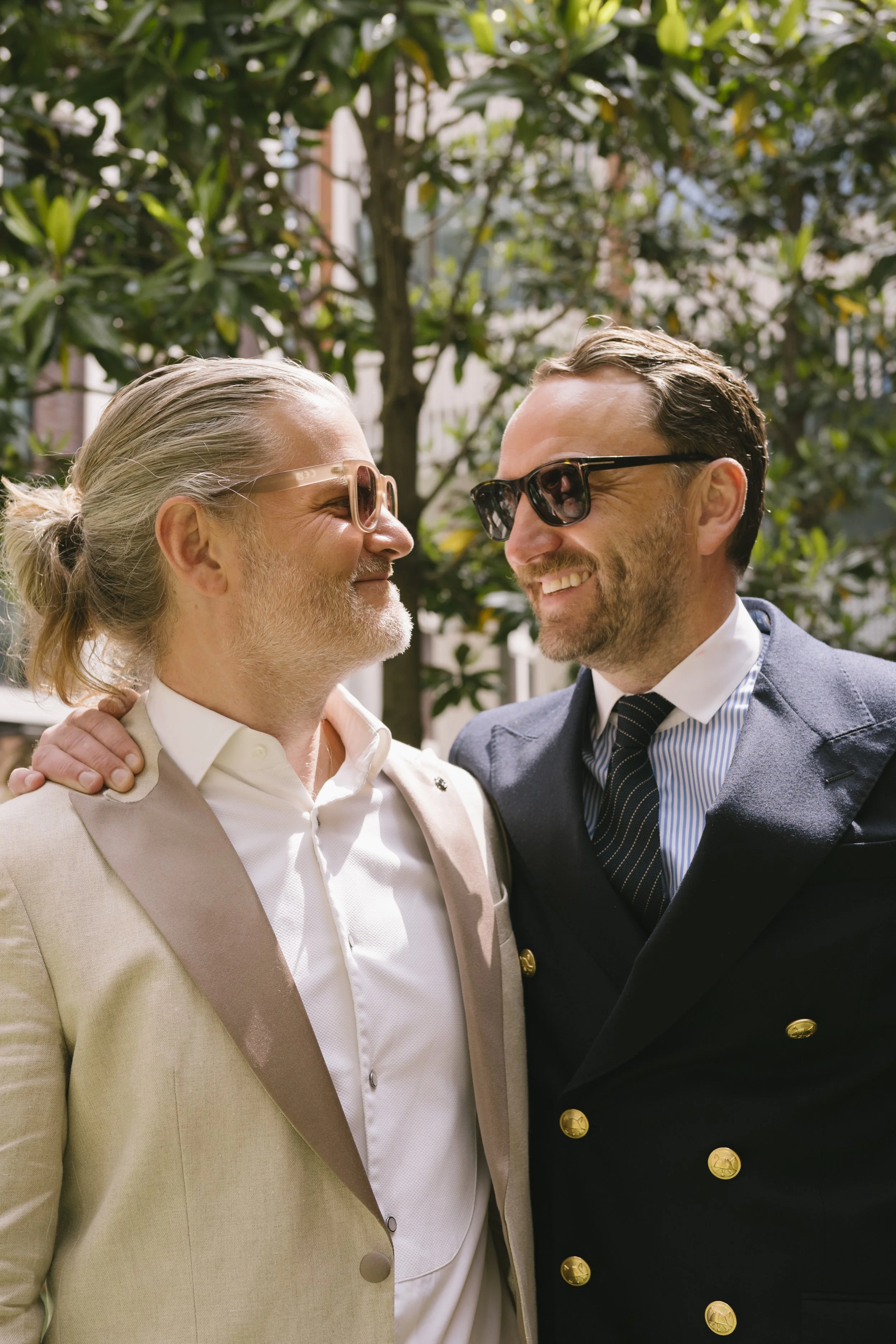 Two grooms share a joyful moment outdoors in soft natural light, captured by Roz Pike Photography. Their stylish suits, sunglasses and relaxed smiles create a warm modern portrait that reflects the love and celebration of a contemporary UK wedding.